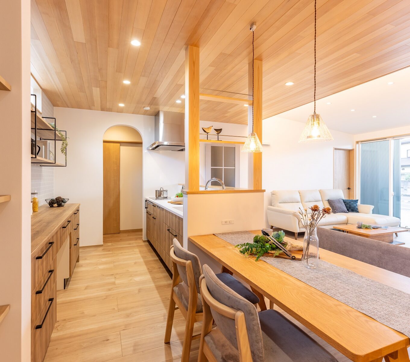 ＬＤＫに入ってまず目を引くのは、キッチン上部の天然木の天井。 キッチンの腰壁から伸びる柱には備え付けの棚を作り、スパイス等を収納できるように。 便利なだけではなく、ナチュラルな温かみのあるアクセントとして作用します。勾配天井の先には大きな窓があり、その先にはドッグランとして遊べる広いお庭と、 それをゆったりと眺められるタイルテラスがあります。